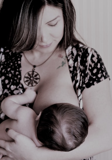 A woman is breastfeeding a baby, holding the child gently in her arms. Her gaze is directed downwards, and she wears a patterned outfit with a large pendant necklace. The scene appears intimate and nurturing.