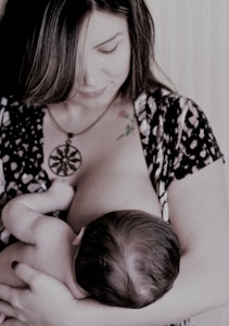 A woman is breastfeeding a baby, holding the child gently in her arms. Her gaze is directed downwards, and she wears a patterned outfit with a large pendant necklace. The scene appears intimate and nurturing.