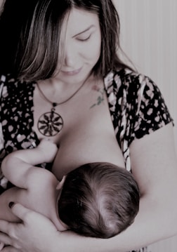 A woman is breastfeeding a baby, holding the child gently in her arms. Her gaze is directed downwards, and she wears a patterned outfit with a large pendant necklace. The scene appears intimate and nurturing.