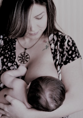 A woman is breastfeeding a baby, holding the child gently in her arms. Her gaze is directed downwards, and she wears a patterned outfit with a large pendant necklace. The scene appears intimate and nurturing.