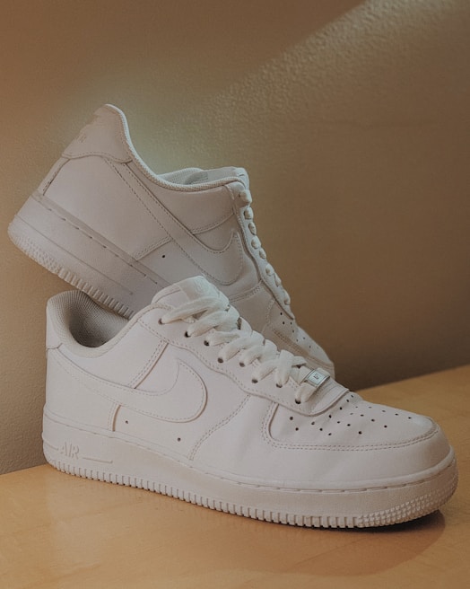 A pair of pristine white sneakers with a classic design are displayed on a wooden surface. The sneakers feature a low-top silhouette with perforations on the toe box and a notable logo on the sides. They are arranged with one shoe leaning against the other, creating a sense of balance and stability.