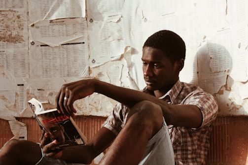 A person is sitting against a wall covered with worn and crumpled papers, deeply focused on reading a book. The room appears to have a rustic and weathered ambiance, and the individual is wearing casual attire.