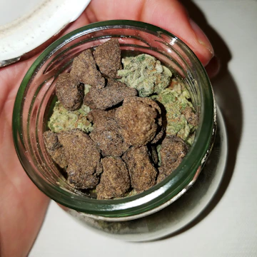 A hand holds a small glass jar filled with irregularly shaped brown and green chunks, possibly a mixture of hashish and cannabis buds.