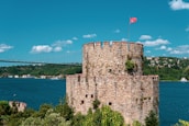 A historic stone fort with crenelated walls is positioned by a large body of water. A red flag with a crescent and star waves atop the fort. A suspension bridge stretches across the background, above green hills dotted with small houses.