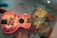 A colorful guitar with western-themed illustrations including cowboys and horses is displayed against a wall. The guitar features a red and yellow color scheme with decorative designs and text. Beside it is a vintage-looking vest with an embroidered pattern, hanging as part of the display.