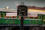 A vintage model train setup featuring a central signpost with directions to Hamburg and Frankfurt. The sign has a black central section with German text and two smaller signs pointing outwards. In the background, there is a green train carriage with a numbered compartment door.