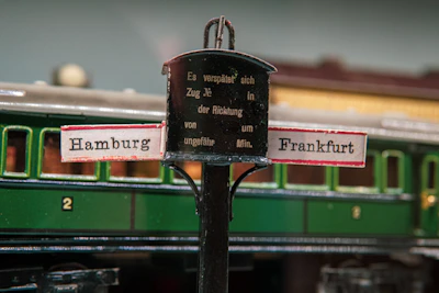 A vintage model train setup featuring a central signpost with directions to Hamburg and Frankfurt. The sign has a black central section with German text and two smaller signs pointing outwards. In the background, there is a green train carriage with a numbered compartment door.
