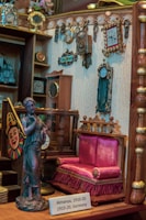 A miniature vintage room with various decorative items from the early 20th century. The setup includes a small vibrant pink upholstered settee with tassels, a detailed statuette of a woman, and several antique clocks and wall hangings. The walls display ornate picture frames and a calendar dated 1910-20. The setting reflects intricate craftsmanship and attention to detail.