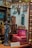 A miniature vintage room with various decorative items from the early 20th century. The setup includes a small vibrant pink upholstered settee with tassels, a detailed statuette of a woman, and several antique clocks and wall hangings. The walls display ornate picture frames and a calendar dated 1910-20. The setting reflects intricate craftsmanship and attention to detail.
