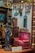 A miniature vintage room with various decorative items from the early 20th century. The setup includes a small vibrant pink upholstered settee with tassels, a detailed statuette of a woman, and several antique clocks and wall hangings. The walls display ornate picture frames and a calendar dated 1910-20. The setting reflects intricate craftsmanship and attention to detail.