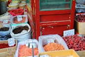 A variety of dried goods displayed in open sacks and boxes at a marketplace. Signs with handwritten prices in Chinese are visible on each sack, indicating different types of dried produce. The setting appears to be an outdoor market, possibly in an Asian country. The collection includes dried fruits, nuts, and seeds, showcasing a colorful assortment.