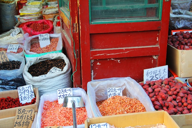 A variety of dried goods displayed in open sacks and boxes at a marketplace. Signs with handwritten prices in Chinese are visible on each sack, indicating different types of dried produce. The setting appears to be an outdoor market, possibly in an Asian country. The collection includes dried fruits, nuts, and seeds, showcasing a colorful assortment.