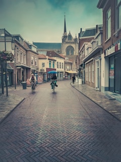 A picturesque street scene with people riding bicycles on a cobblestone path. Historic European-style buildings with a church spire in the background create a serene atmosphere. The street is lined with quaint shops and flower pots, under a slightly overcast sky.