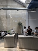 A modern coffee shop with industrial design elements, including exposed ductwork and textured brick walls. The space features a long counter where baristas are preparing drinks, and there are several people sitting and chatting along a bench. Indoor plants and sleek lighting contribute to a minimalist aesthetic.