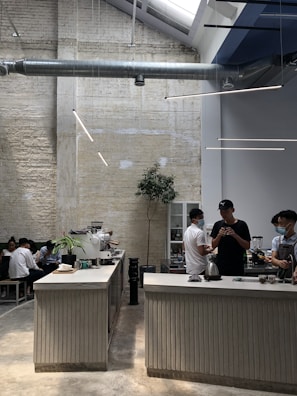 A modern coffee shop with industrial design elements, including exposed ductwork and textured brick walls. The space features a long counter where baristas are preparing drinks, and there are several people sitting and chatting along a bench. Indoor plants and sleek lighting contribute to a minimalist aesthetic.