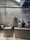 A modern coffee shop with industrial design elements, including exposed ductwork and textured brick walls. The space features a long counter where baristas are preparing drinks, and there are several people sitting and chatting along a bench. Indoor plants and sleek lighting contribute to a minimalist aesthetic.