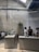 A modern coffee shop with industrial design elements, including exposed ductwork and textured brick walls. The space features a long counter where baristas are preparing drinks, and there are several people sitting and chatting along a bench. Indoor plants and sleek lighting contribute to a minimalist aesthetic.