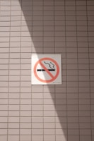 A no smoking sign is displayed on a wall with a light gray tile pattern. The sign features a cigarette symbol crossed out with a red circle and diagonal line. Sunlight casts a shadow across part of the tiles and the sign, creating contrasting areas of light and shadow.