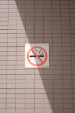 A no smoking sign is displayed on a wall with a light gray tile pattern. The sign features a cigarette symbol crossed out with a red circle and diagonal line. Sunlight casts a shadow across part of the tiles and the sign, creating contrasting areas of light and shadow.