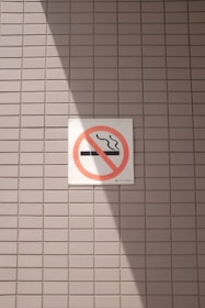 A no smoking sign is displayed on a wall with a light gray tile pattern. The sign features a cigarette symbol crossed out with a red circle and diagonal line. Sunlight casts a shadow across part of the tiles and the sign, creating contrasting areas of light and shadow.