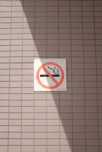 A no smoking sign is displayed on a wall with a light gray tile pattern. The sign features a cigarette symbol crossed out with a red circle and diagonal line. Sunlight casts a shadow across part of the tiles and the sign, creating contrasting areas of light and shadow.