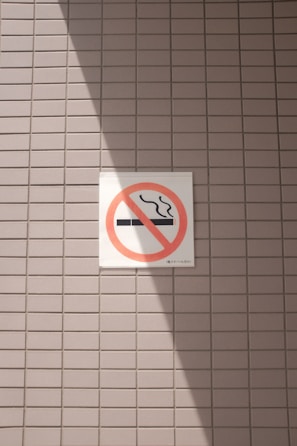 A no smoking sign is displayed on a wall with a light gray tile pattern. The sign features a cigarette symbol crossed out with a red circle and diagonal line. Sunlight casts a shadow across part of the tiles and the sign, creating contrasting areas of light and shadow.
