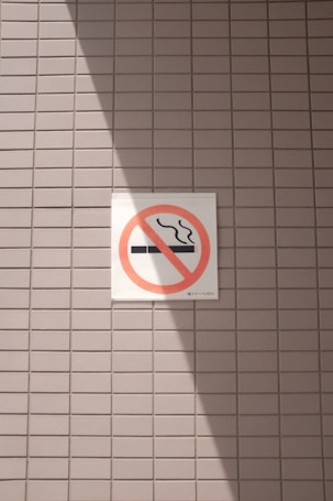 A no smoking sign is displayed on a wall with a light gray tile pattern. The sign features a cigarette symbol crossed out with a red circle and diagonal line. Sunlight casts a shadow across part of the tiles and the sign, creating contrasting areas of light and shadow.
