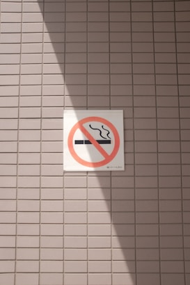 A no smoking sign is displayed on a wall with a light gray tile pattern. The sign features a cigarette symbol crossed out with a red circle and diagonal line. Sunlight casts a shadow across part of the tiles and the sign, creating contrasting areas of light and shadow.