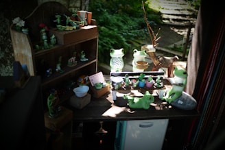 A collection of cute frog figurines is arranged on a small wooden shelf and a table. Some frogs are holding objects like a cup and a watering can, while others sit peacefully. The scene is set in a cozy, shaded area with plants in the background, creating a whimsical atmosphere.
