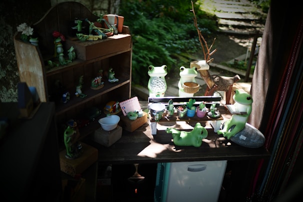 A collection of cute frog figurines is arranged on a small wooden shelf and a table. Some frogs are holding objects like a cup and a watering can, while others sit peacefully. The scene is set in a cozy, shaded area with plants in the background, creating a whimsical atmosphere.