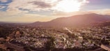 A panoramic view of a suburban neighborhood nestled in a valley with a backdrop of rolling hills. The scene is bathed in the warm, golden light of the setting sun, casting long shadows and creating a peaceful, serene atmosphere. Houses are scattered amidst lush green trees, with curving roads weaving through the community.
