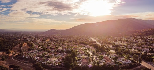 A panoramic view of a suburban neighborhood nestled in a valley with a backdrop of rolling hills. The scene is bathed in the warm, golden light of the setting sun, casting long shadows and creating a peaceful, serene atmosphere. Houses are scattered amidst lush green trees, with curving roads weaving through the community.