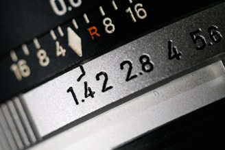 Close-up view of a camera lens aperture scale, displaying various f-stop numbers such as 1.4, 2.8, and 5.6. The R marking and a series of numerical indicators provide exposure settings.