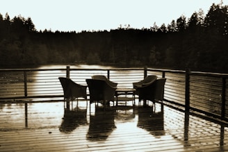 A wooden deck with a set of woven chairs and a small table overlooks a serene body of water surrounded by dense, dark trees. The surface of the deck appears damp, reflecting the furniture and creating a calm, tranquil scene.
