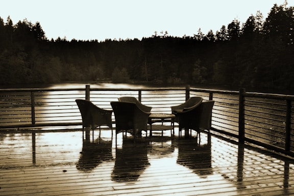 A wooden deck with a set of woven chairs and a small table overlooks a serene body of water surrounded by dense, dark trees. The surface of the deck appears damp, reflecting the furniture and creating a calm, tranquil scene.