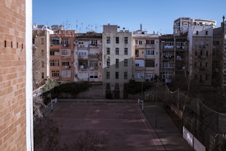 A row of older, tightly packed apartment buildings with varied colored facades face a vacant basketball court. The architecture features multiple balconies, some with hanging clothes, and is surrounded by a boundary fence. In the foreground, bare trees stand under clear blue skies.
