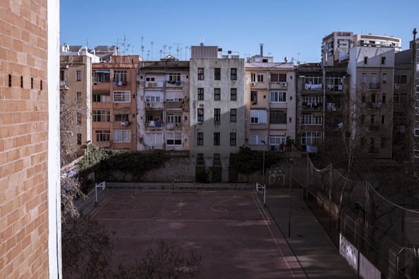 A row of older, tightly packed apartment buildings with varied colored facades face a vacant basketball court. The architecture features multiple balconies, some with hanging clothes, and is surrounded by a boundary fence. In the foreground, bare trees stand under clear blue skies.