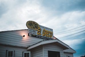 A pizza restaurant sign sits atop a white building with wooden siding. The sign, decorated with an image of a pizza, displays the text 'EL GRECO - The Best Pizza in Town' alongside three phone numbers. Overcast skies and power lines form the background, casting a moody atmosphere.