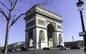 The image features a large neoclassical monument with ornate sculptures and a massive archway, set against a clear blue sky. Surrounding the monument are streets bustling with cars. In the background, a partially visible iconic tower rises above a tree-lined area. Leafless trees frame the foreground, while a streetlamp adds an urban touch. A street sign is visible, indicating 'Avenue Carnot'.
