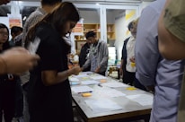 A group of people are gathered around a table covered with papers, pens, and sticky notes. They appear to be engaged in a collaborative activity or workshop, focused and discussing ideas. The setting suggests an indoor environment with shelves and posters in the background.