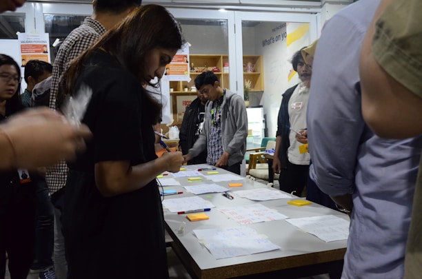A group of people are gathered around a table covered with papers, pens, and sticky notes. They appear to be engaged in a collaborative activity or workshop, focused and discussing ideas. The setting suggests an indoor environment with shelves and posters in the background.