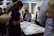 A group of people are gathered around a table covered with papers, pens, and sticky notes. They appear to be engaged in a collaborative activity or workshop, focused and discussing ideas. The setting suggests an indoor environment with shelves and posters in the background.