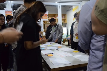 A group of people are gathered around a table covered with papers, pens, and sticky notes. They appear to be engaged in a collaborative activity or workshop, focused and discussing ideas. The setting suggests an indoor environment with shelves and posters in the background.