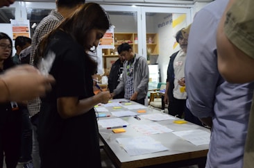A group of people are gathered around a table covered with papers, pens, and sticky notes. They appear to be engaged in a collaborative activity or workshop, focused and discussing ideas. The setting suggests an indoor environment with shelves and posters in the background.