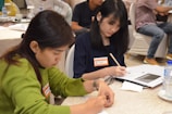 Two people are seated at a table, focused on writing in notebooks. The woman on the left is wearing a green sweater and is concentrating on her work. The woman on the right has dark hair and is wearing a dark shirt, also engaged in writing. There are name tags on their clothing. The setting appears to be a meeting or workshop with other attendees in the background.