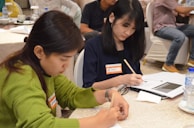 Two people are seated at a table, focused on writing in notebooks. The woman on the left is wearing a green sweater and is concentrating on her work. The woman on the right has dark hair and is wearing a dark shirt, also engaged in writing. There are name tags on their clothing. The setting appears to be a meeting or workshop with other attendees in the background.