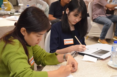 Two people are seated at a table, focused on writing in notebooks. The woman on the left is wearing a green sweater and is concentrating on her work. The woman on the right has dark hair and is wearing a dark shirt, also engaged in writing. There are name tags on their clothing. The setting appears to be a meeting or workshop with other attendees in the background.
