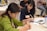 Two people are seated at a table, focused on writing in notebooks. The woman on the left is wearing a green sweater and is concentrating on her work. The woman on the right has dark hair and is wearing a dark shirt, also engaged in writing. There are name tags on their clothing. The setting appears to be a meeting or workshop with other attendees in the background.