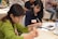 Two people are seated at a table, focused on writing in notebooks. The woman on the left is wearing a green sweater and is concentrating on her work. The woman on the right has dark hair and is wearing a dark shirt, also engaged in writing. There are name tags on their clothing. The setting appears to be a meeting or workshop with other attendees in the background.