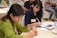 Two people are seated at a table, focused on writing in notebooks. The woman on the left is wearing a green sweater and is concentrating on her work. The woman on the right has dark hair and is wearing a dark shirt, also engaged in writing. There are name tags on their clothing. The setting appears to be a meeting or workshop with other attendees in the background.